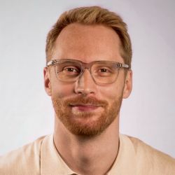 Professional headshot of Tom Linford-Grayson, Digital Marketing Partner, wearing clear-framed glasses and a beige polo shirt.